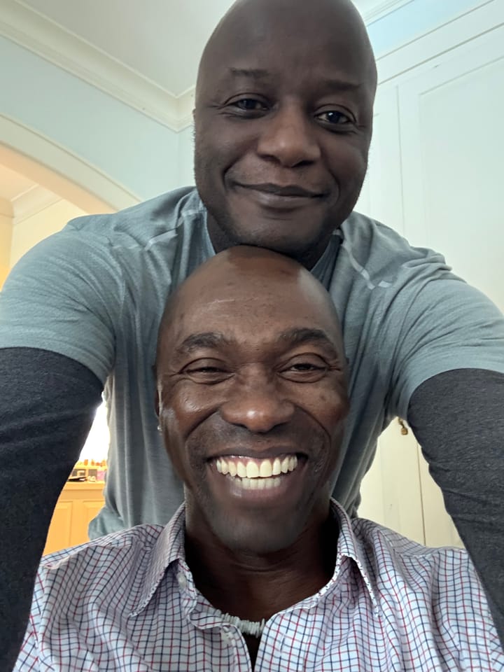 Your Hero and his friend smiling for the camera in a close-up selfie. My friend has a broad smile and is wearing a checkered shirt and YH is leaning in, looking happy. We are indoors